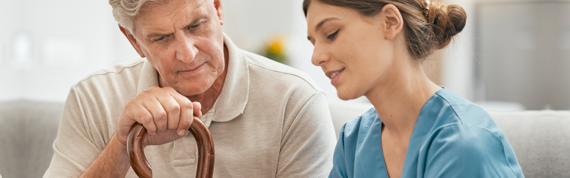 Elderly man holding a cane sits next to a smiling nurse in blue scrubs, both engaged in conversation, conveying care and support in a home setting.