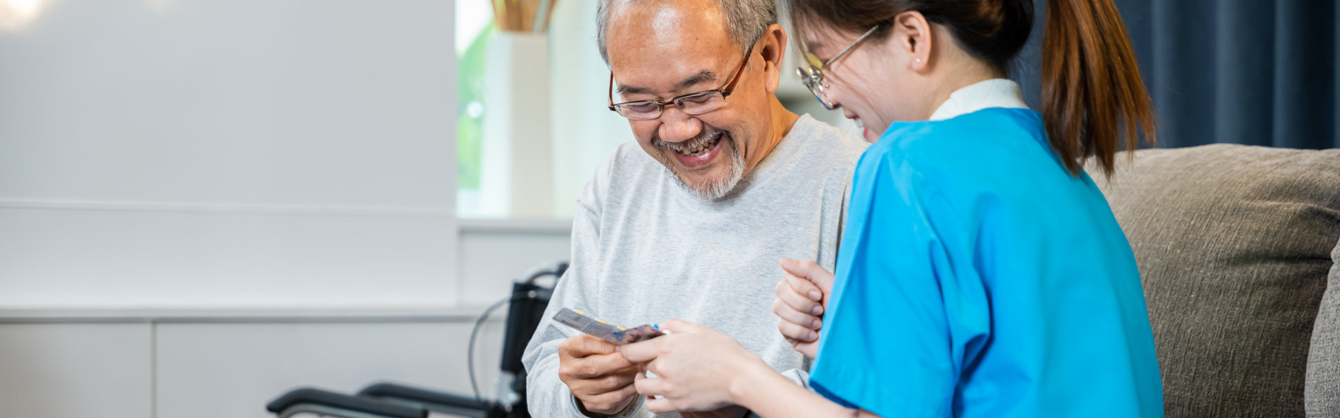 A smiling elderly manr holds cards while chatting with a smiling nurse in blue scrubs in a cozy, warmly lit room.